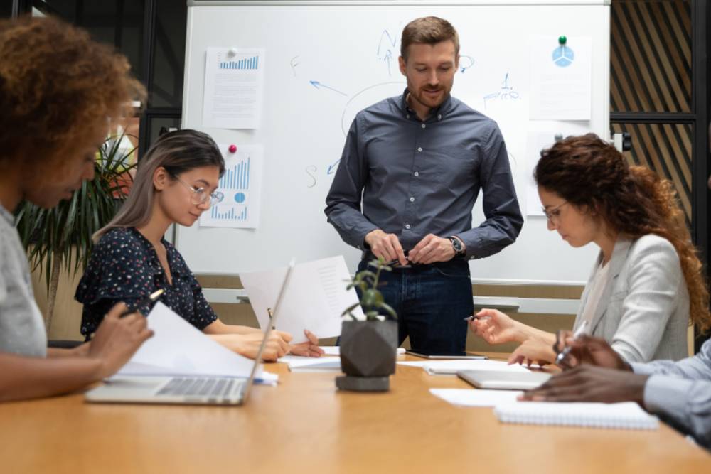 Multiethnic staff take part in educational training led by boss or coach participants do task writing solutions seated at desk in boardroom