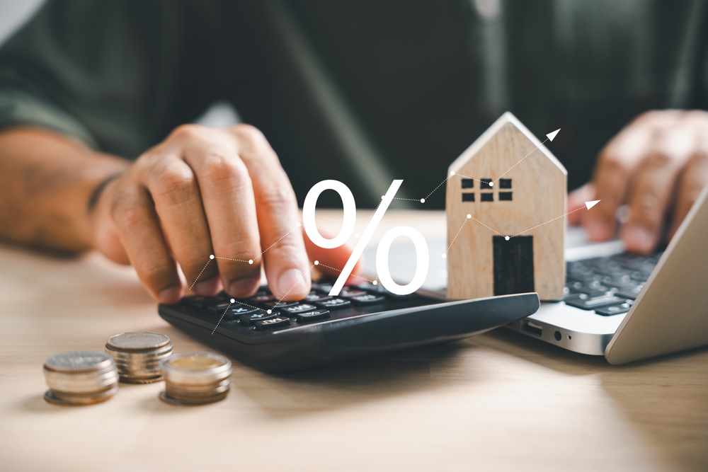 Hand on calculators, strategizing home refinance. Wooden house model, buy and rent note on desk.